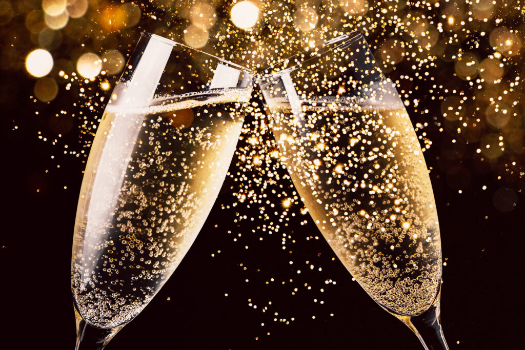 Two glasses of champagne cheerfully clink against a dark background illuminated by golden sparkles, evoking a festive New Year’s celebration.