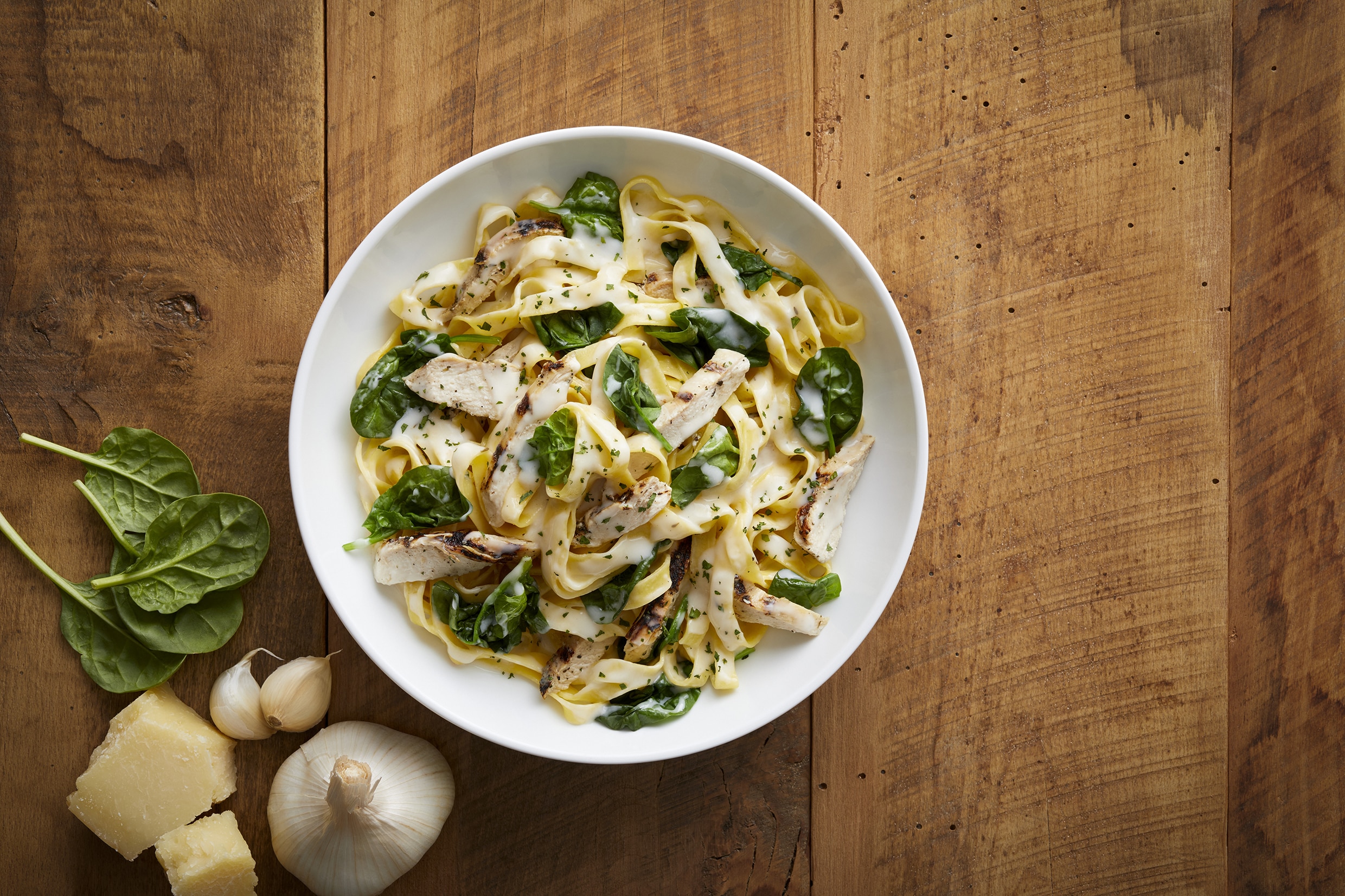 Bravo's Pasta Woozie in a white bowl resting on a wooden table next to garlic, parmesean, and spinach leaves.